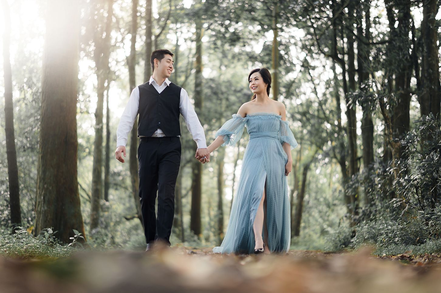 Couple holding hands walking through sunlit forest, woman in blue off-shoulder gown, man in black vest and trousers.
