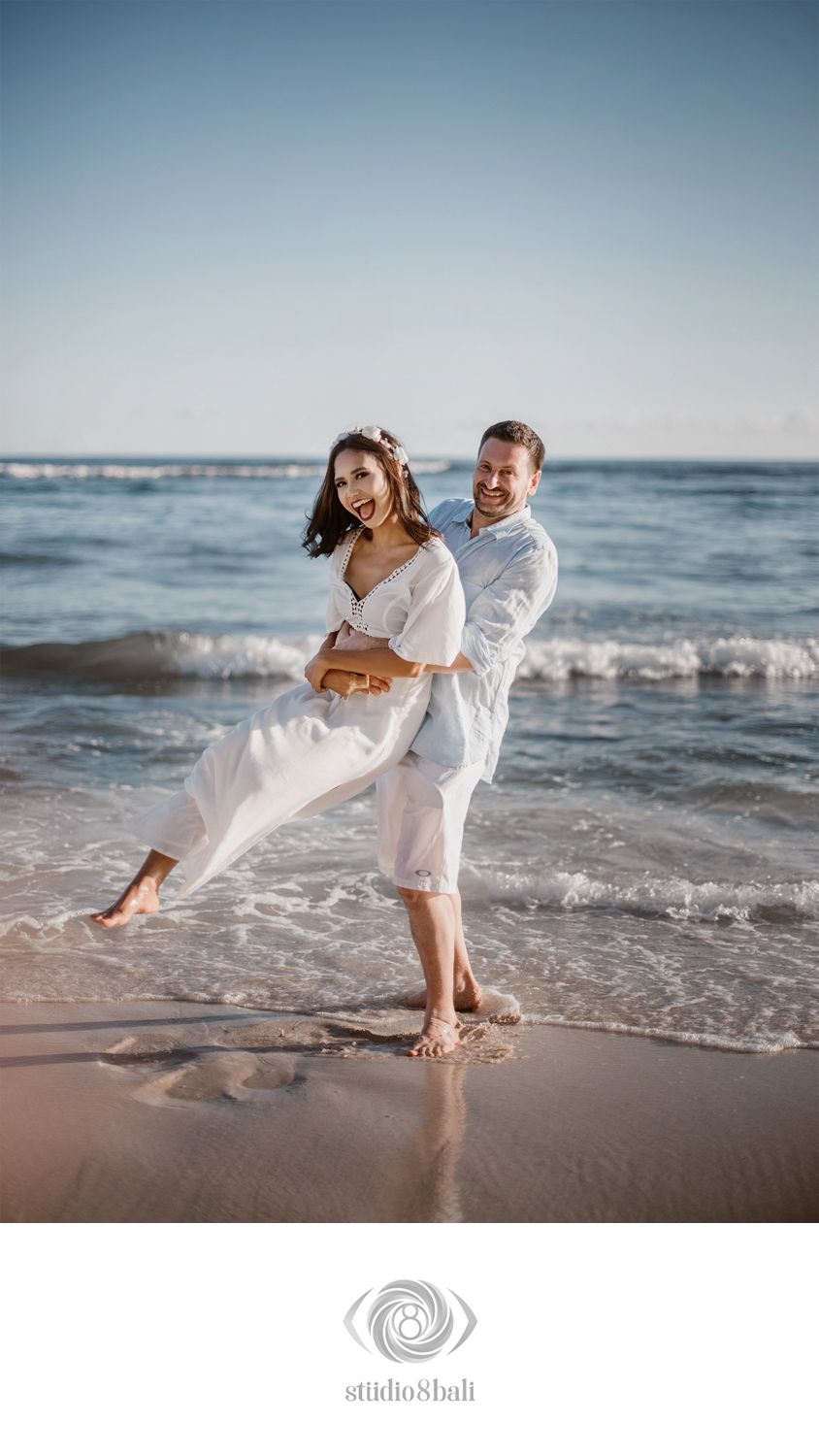 Joyful couple in white outfits posing on a sandy beach with ocean waves, captured by Studio 8 Bali.
