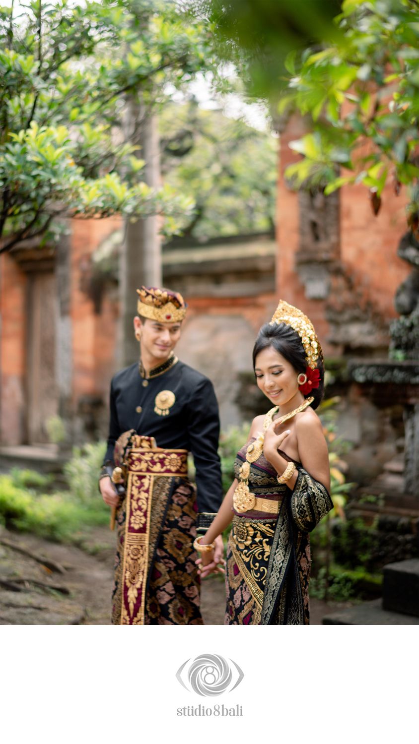 Balinese couple in traditional gold and black ceremonial wedding attire posing in a lush temple garden in Bali.
