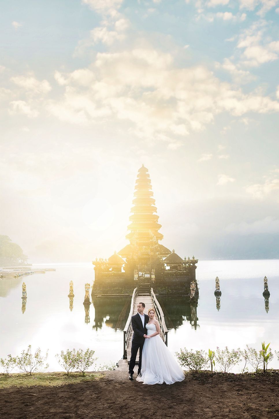 Bride and groom pose before Pura Ulun Danu Bratan temple on Lake Beratan in Bali at golden sunrise.