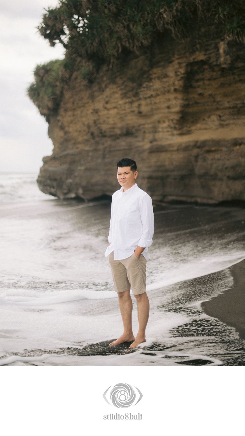 Man in white shirt and khaki shorts standing barefoot on a black sand beach in Bali with rocky cliffs behind.
