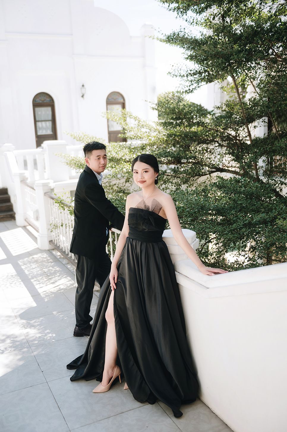 Couple in matching black formal wear posing on a sunlit white balcony with lush green trees in the background.