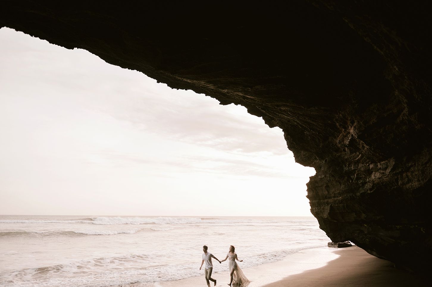 Couple holding hands on sandy beach inside dramatic sea cave arch with ocean waves and glowing sky.