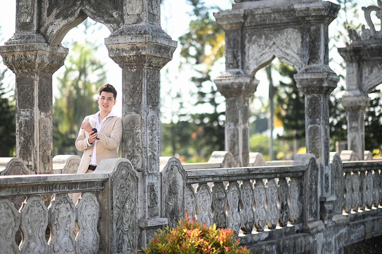 Man in beige suit holding phone among ornate stone columns and balustrade in a lush garden setting.
