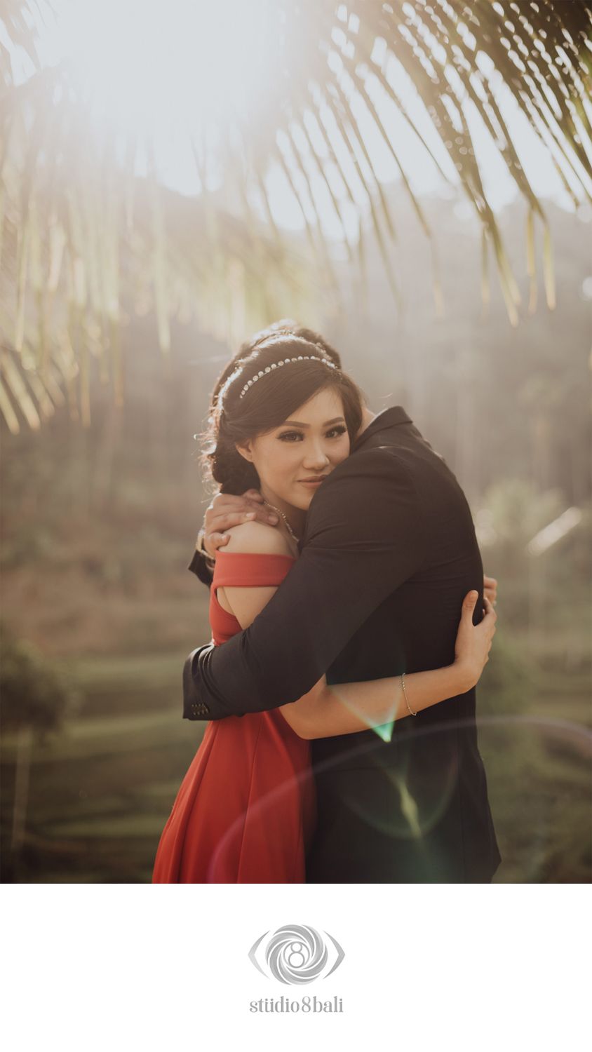 Couple embracing romantically outdoors in Bali, woman in red off-shoulder dress with pearl headband, soft golden sunlight.