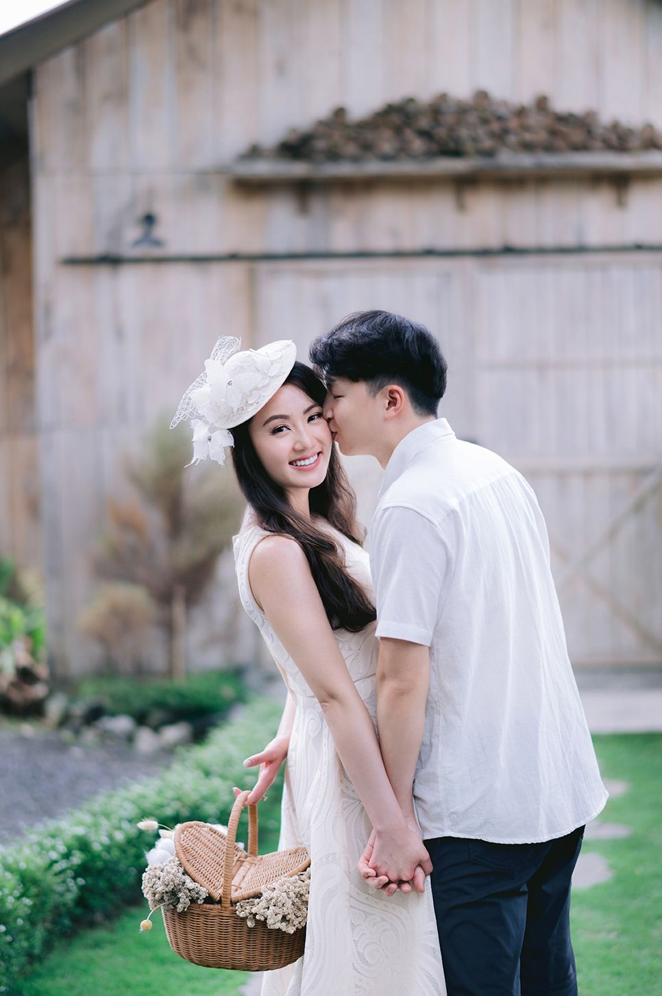 A smiling woman in a white dress and fascinator holds a wicker basket while her partner kisses her cheek outdoors.