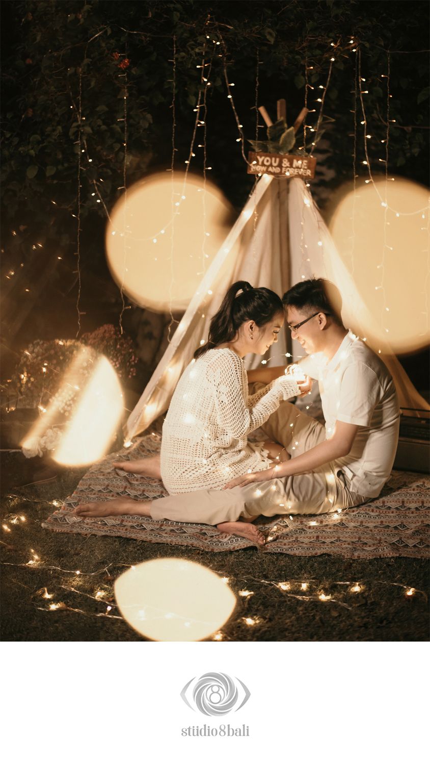 Romantic couple sitting together under a glowing teepee tent surrounded by fairy lights at night.