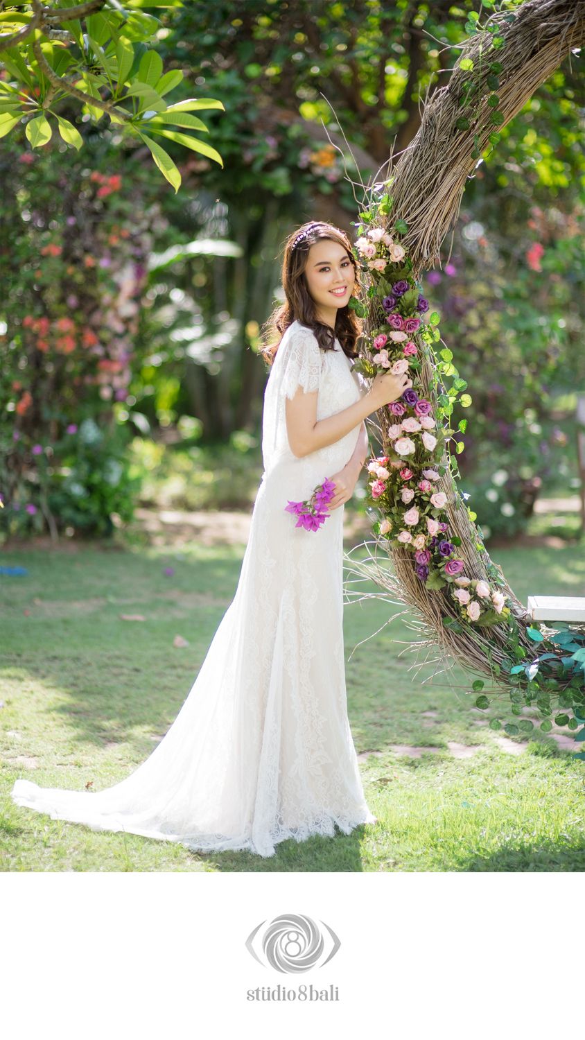 Bride in white lace gown poses beside a floral-adorned tree in a lush garden, smiling elegantly.
