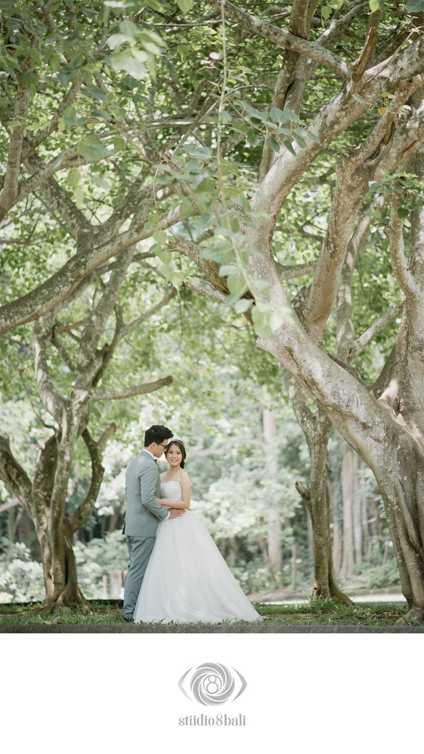 A bride and groom embrace under a canopy of lush green trees in a romantic outdoor wedding portrait.