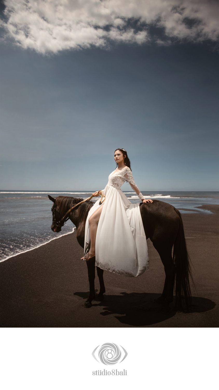 A bride in a white gown rides a dark horse on a black sand beach under a dramatic cloudy sky in Bali.