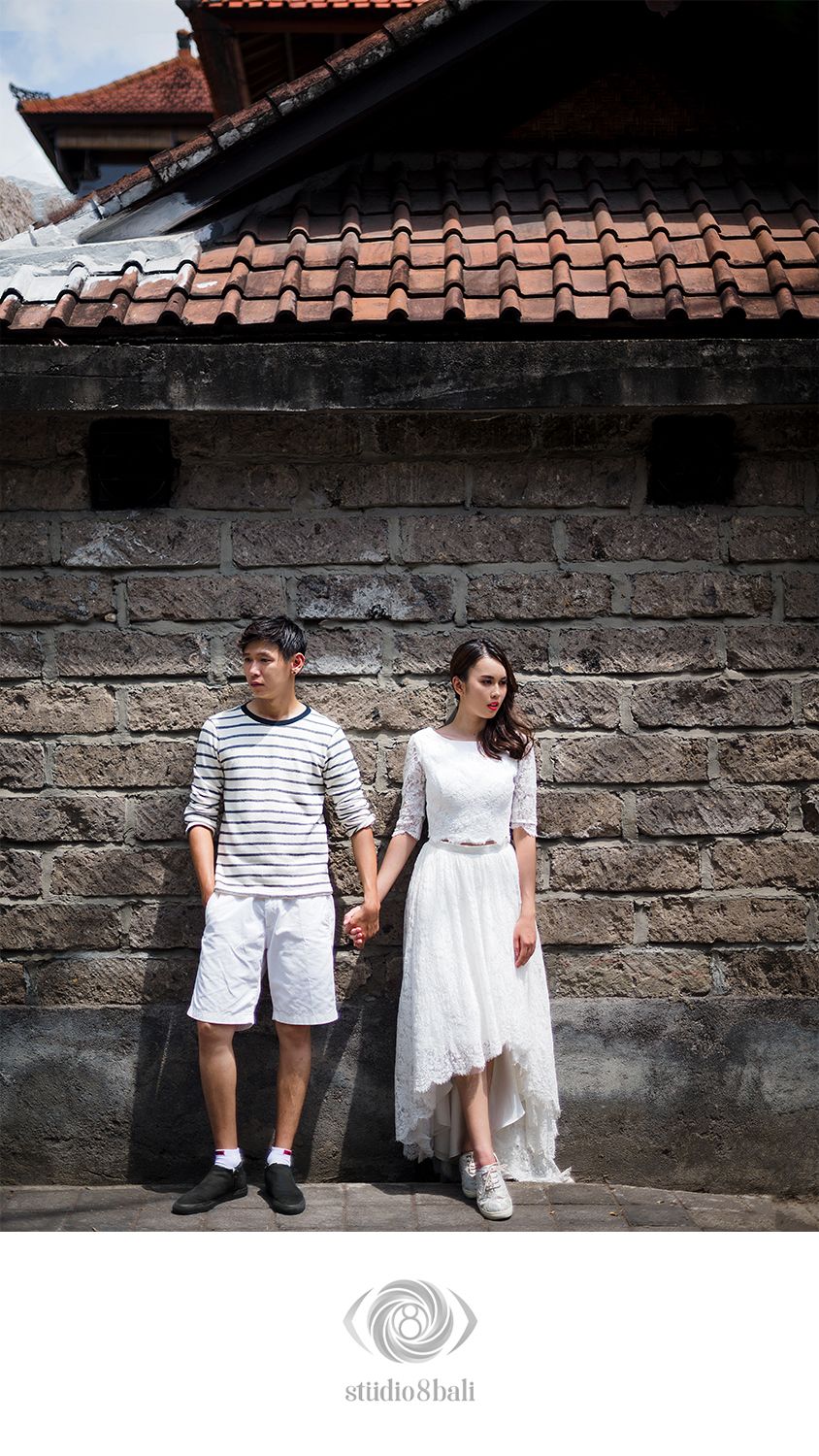 Couple holding hands against a stone wall, man in striped shirt and shorts, woman in elegant white lace dress, Bali.