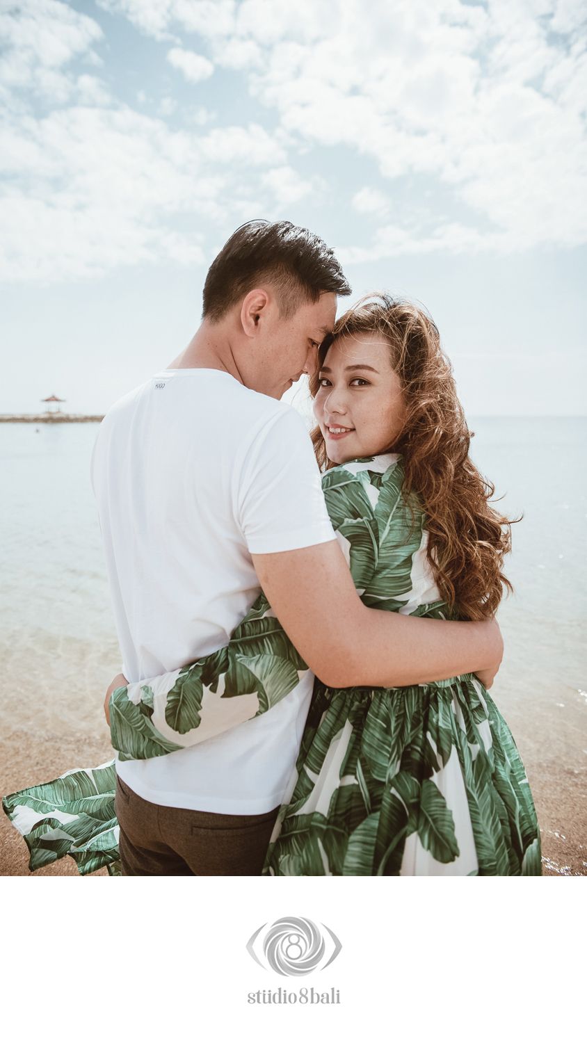Romantic couple embracing on a beach in Bali, woman in tropical leaf print dress, man in white shirt, ocean backdrop.