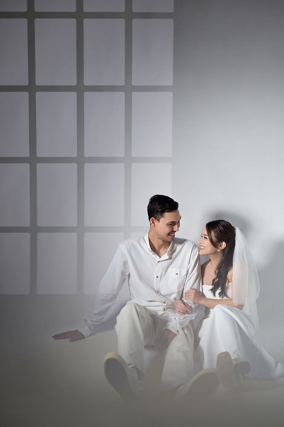 A couple in white wedding attire sits together, smiling at each other against a softly lit grid-patterned backdrop.