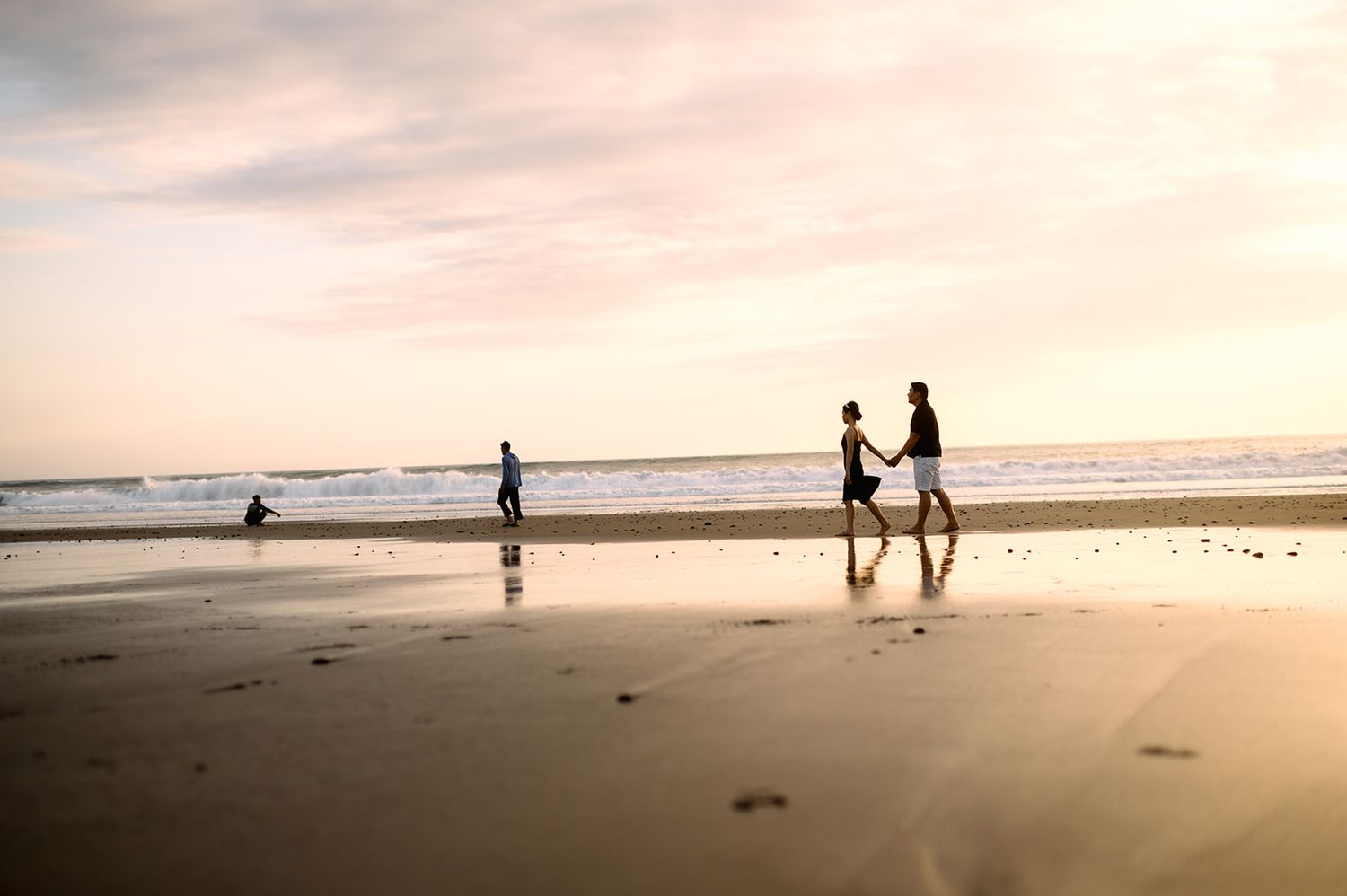 Couple walking hand in hand on a reflective beach at sunset with ocean waves.