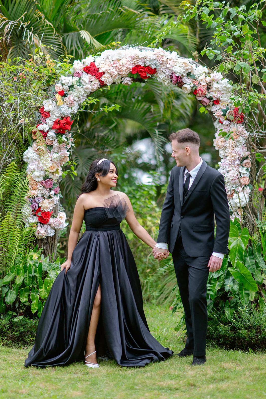 Couple in black formalwear holding hands under a floral arch in a lush garden setting.