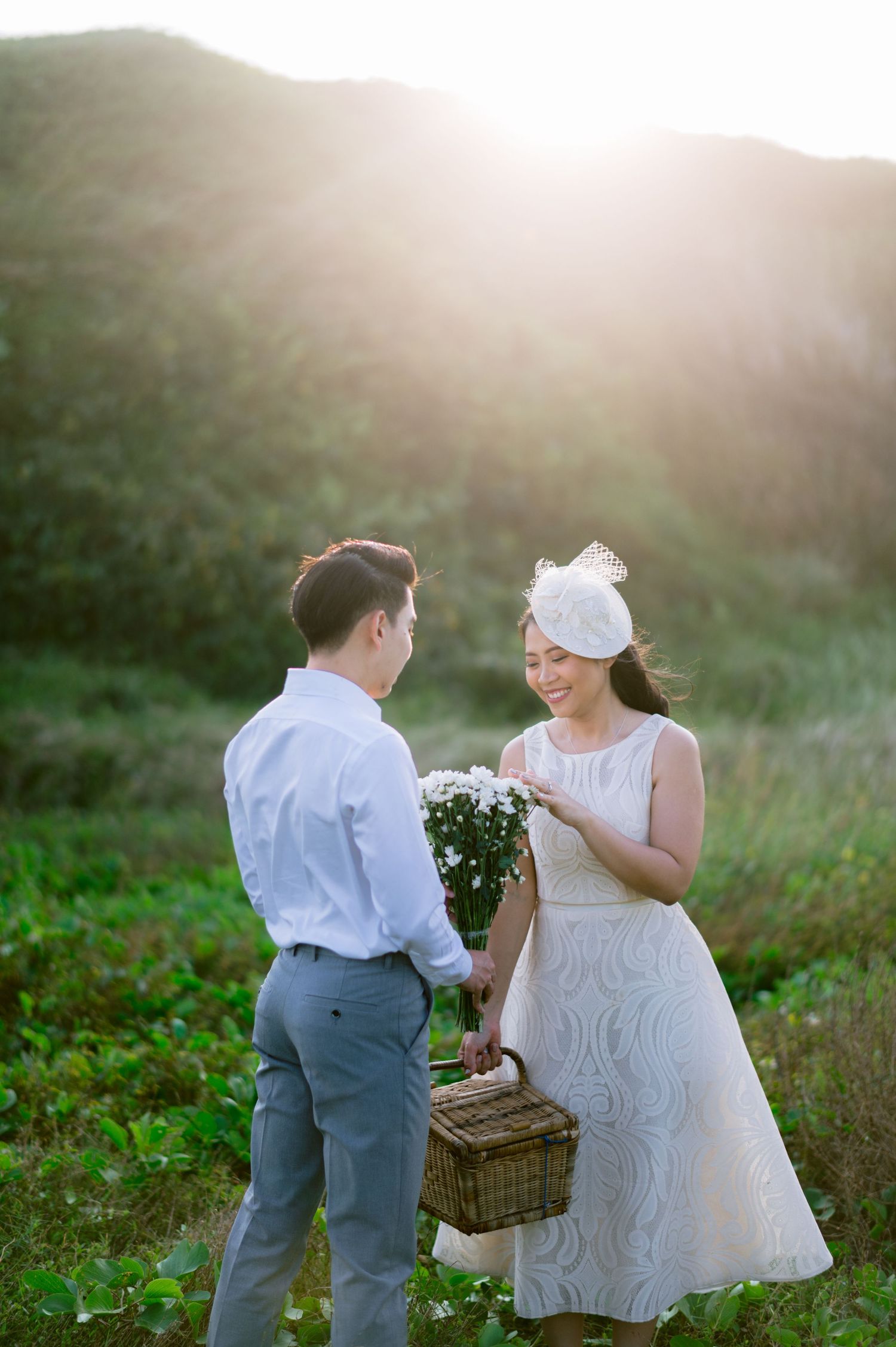 Couple in elegant attire exchange flowers at golden hour in a lush green field, woman in white dress holding picnic basket.