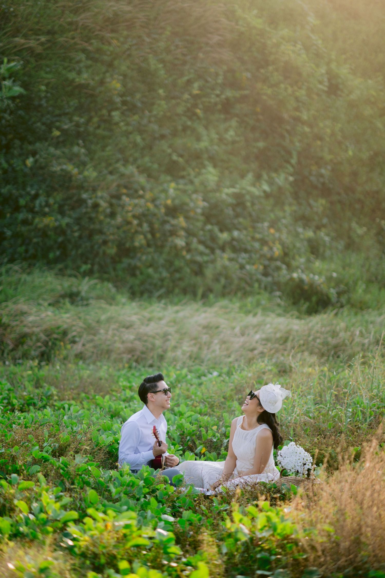 Couple in white outfits sitting in a lush green meadow, sharing a romantic moment during a sunny outdoor photo session.