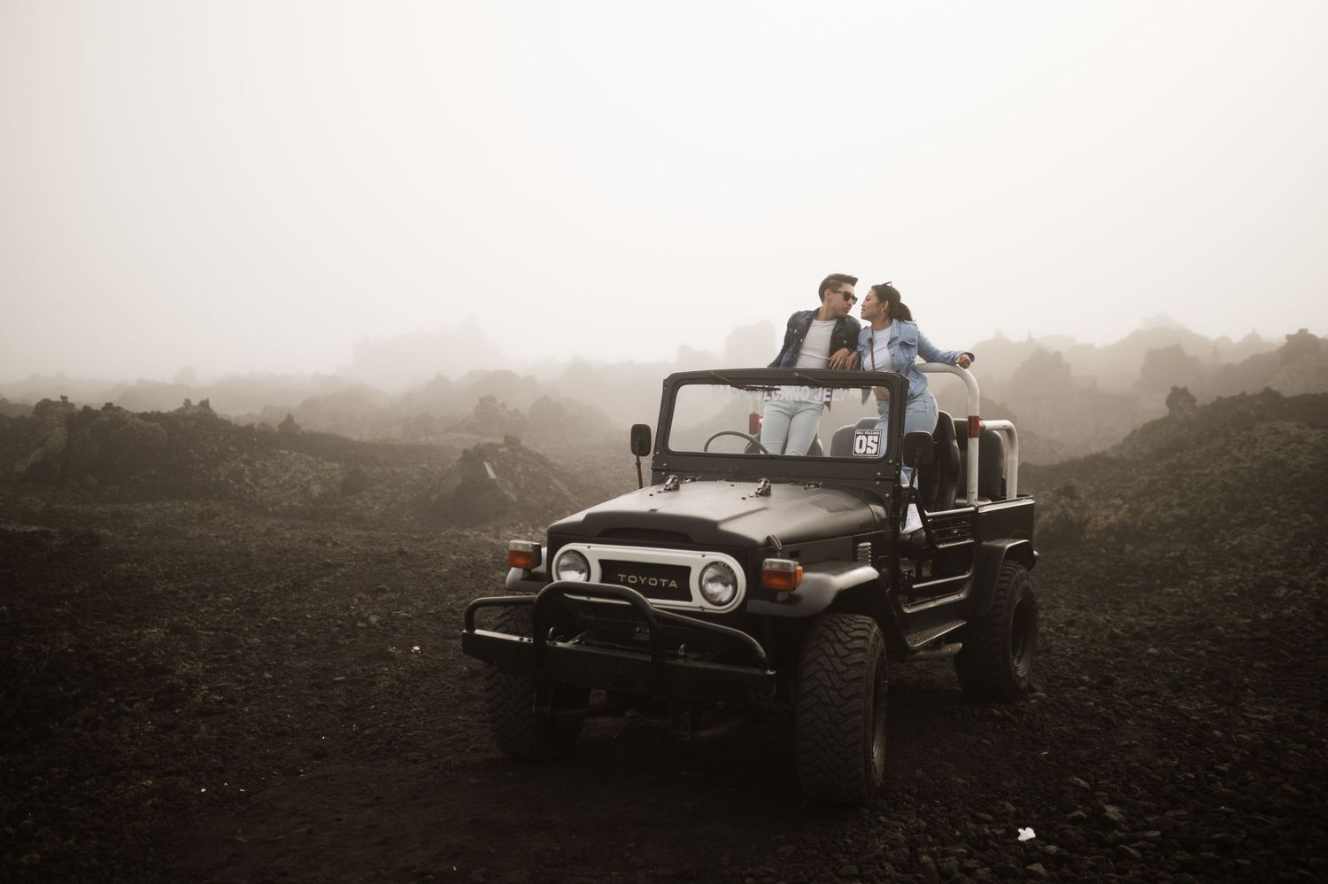 Couple standing on a vintage black jeep in a misty volcanic landscape during a foggy adventure trip.