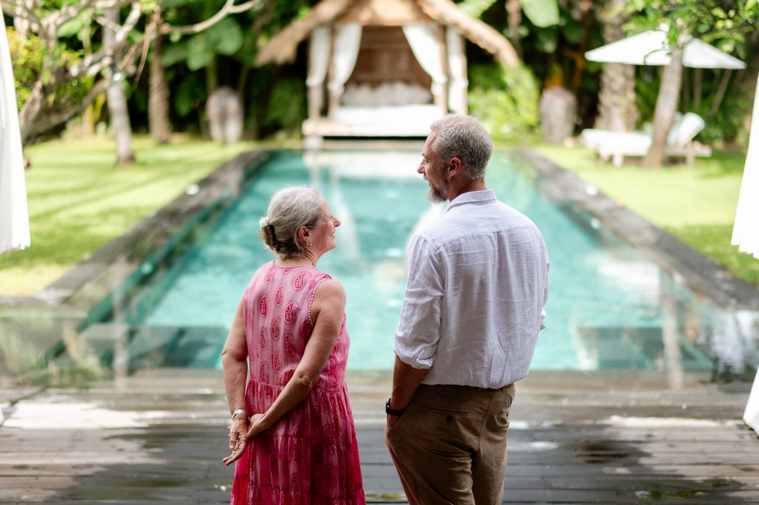 Elderly couple standing by a tropical villa pool surrounded by lush greenery, sharing a conversation outdoors.