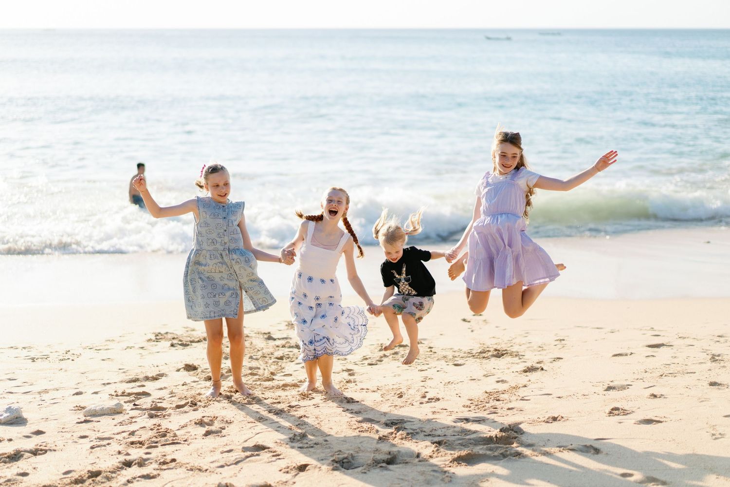 Four children joyfully jumping and running on a sunny sandy beach with ocean waves in the background.