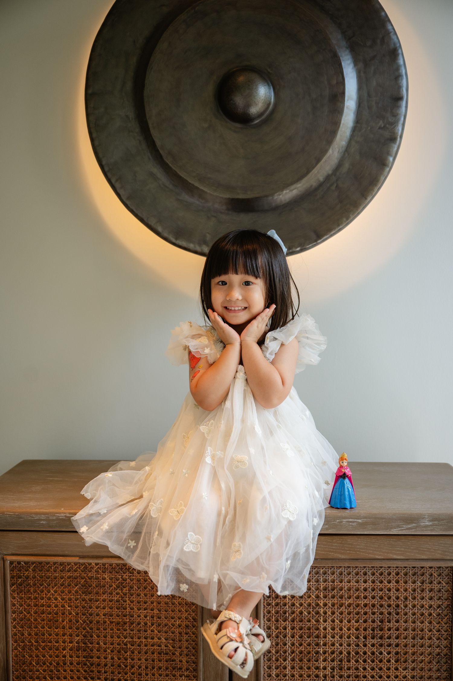 A young girl in a white tulle dress poses cheerfully on a wooden cabinet in front of a large decorative gong.