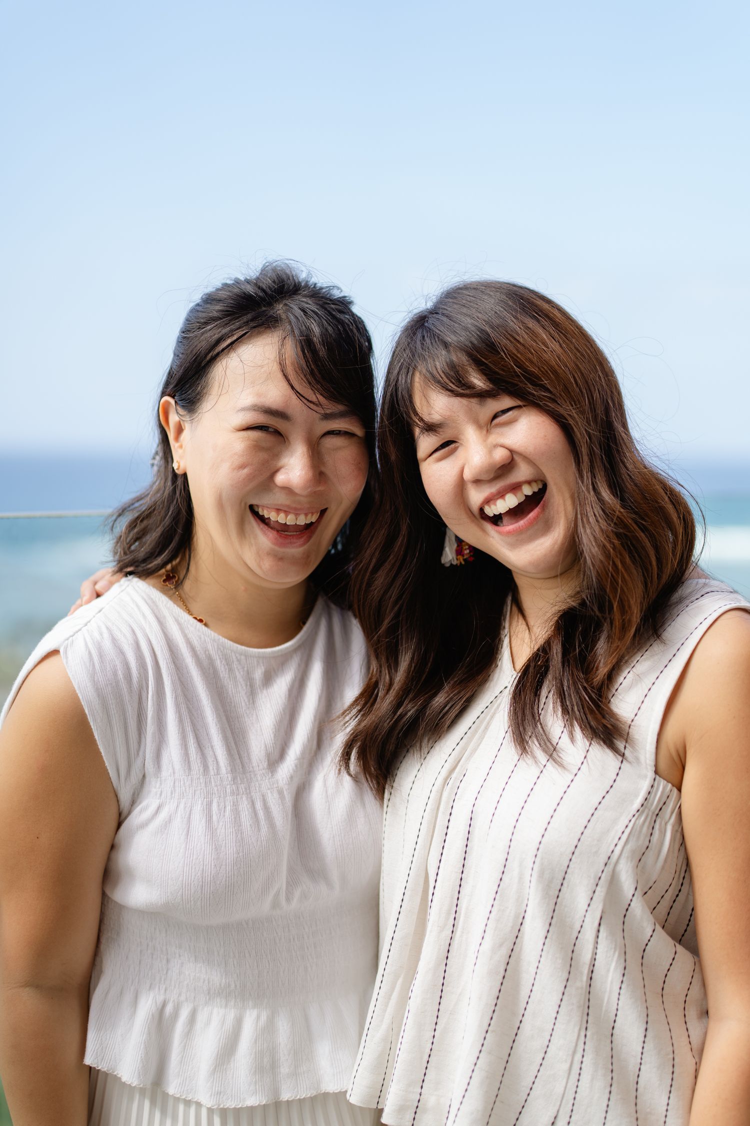 Two young Asian women laughing joyfully together outdoors with ocean and blue sky in background, wearing white sleeveless tops.