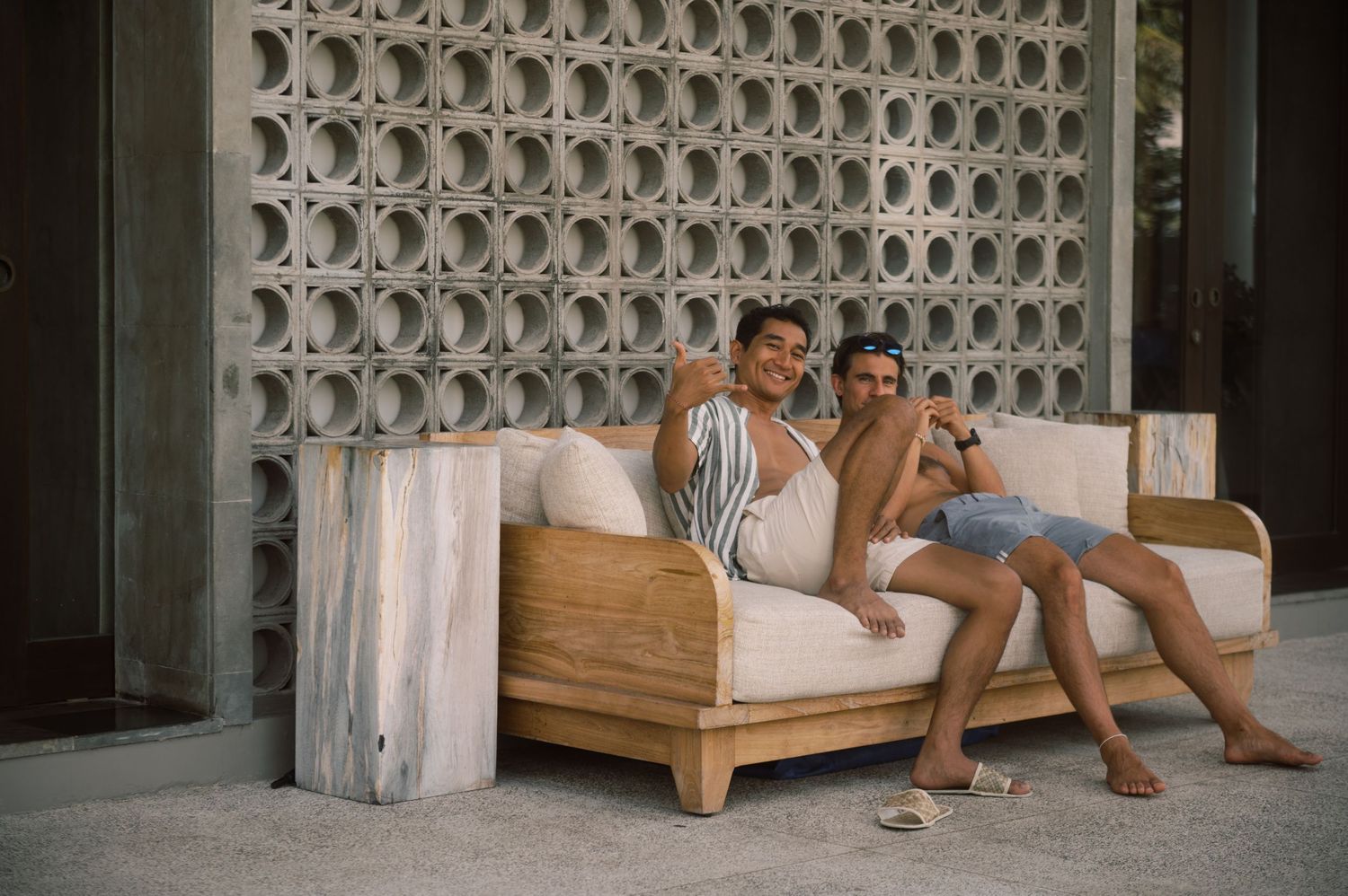 Two people relaxing on a wooden outdoor sofa against a decorative concrete block wall.