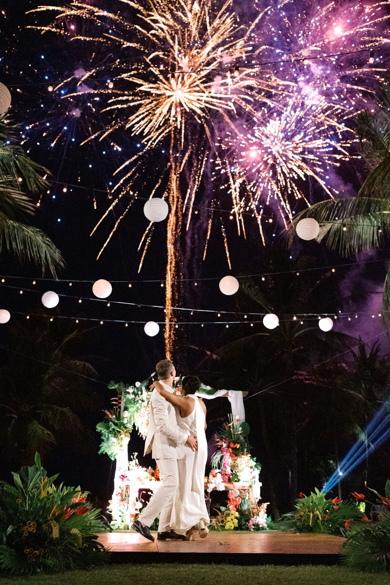 Couple shares first dance under spectacular fireworks display at tropical outdoor wedding reception at night.