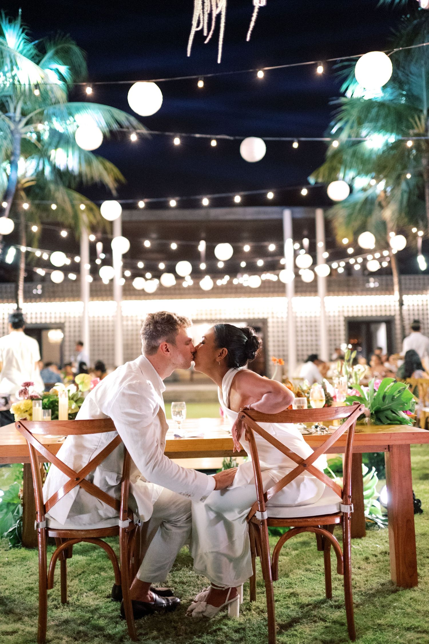 Couple sharing a kiss at an outdoor wedding reception at night, surrounded by string lights and palm trees.