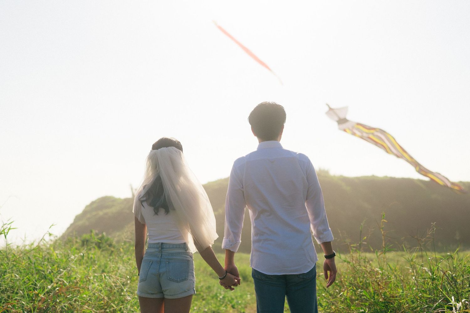 Couple holding hands in a green field watching a kite fly, with hills and bright sky in the background.