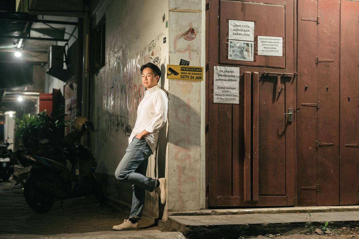 Man in white shirt and jeans leaning against a weathered wall at night on a quiet urban street.