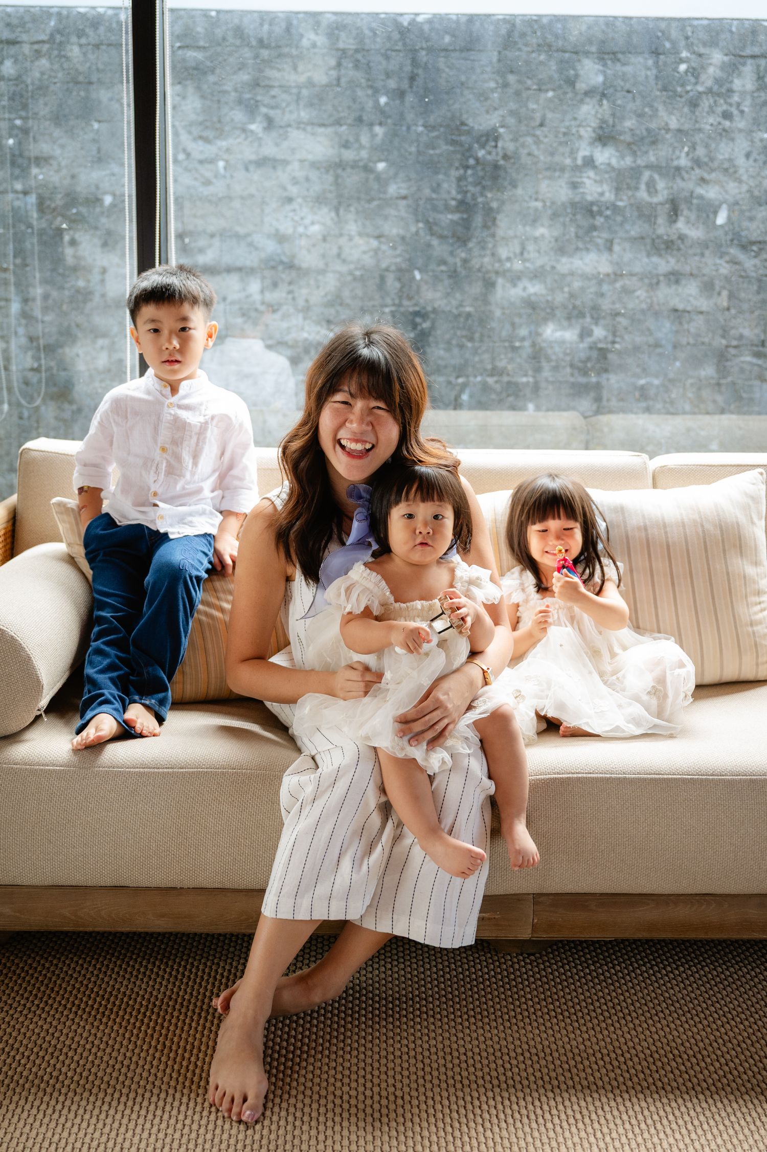 Smiling mother sitting on beige sofa with three young children, enjoying a warm family moment near large windows.