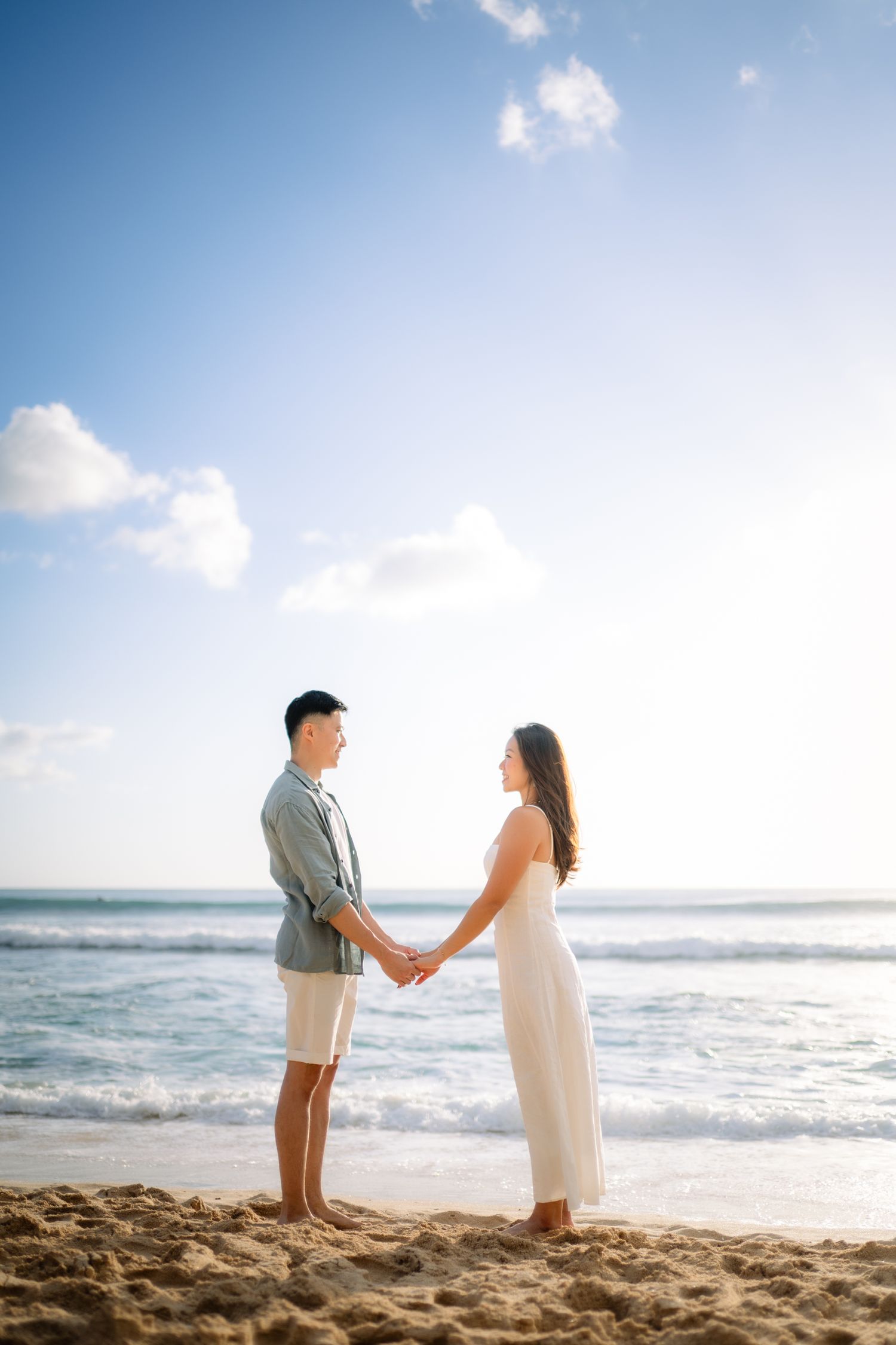Couple holding hands on a sunny beach with ocean waves, wearing casual white dress and gray shirt.