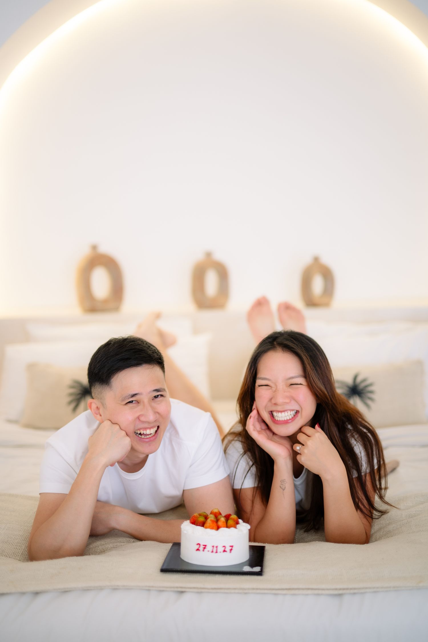 Happy Asian couple lying on bed with anniversary cake dated 27.11.17, smiling joyfully in white bedroom.