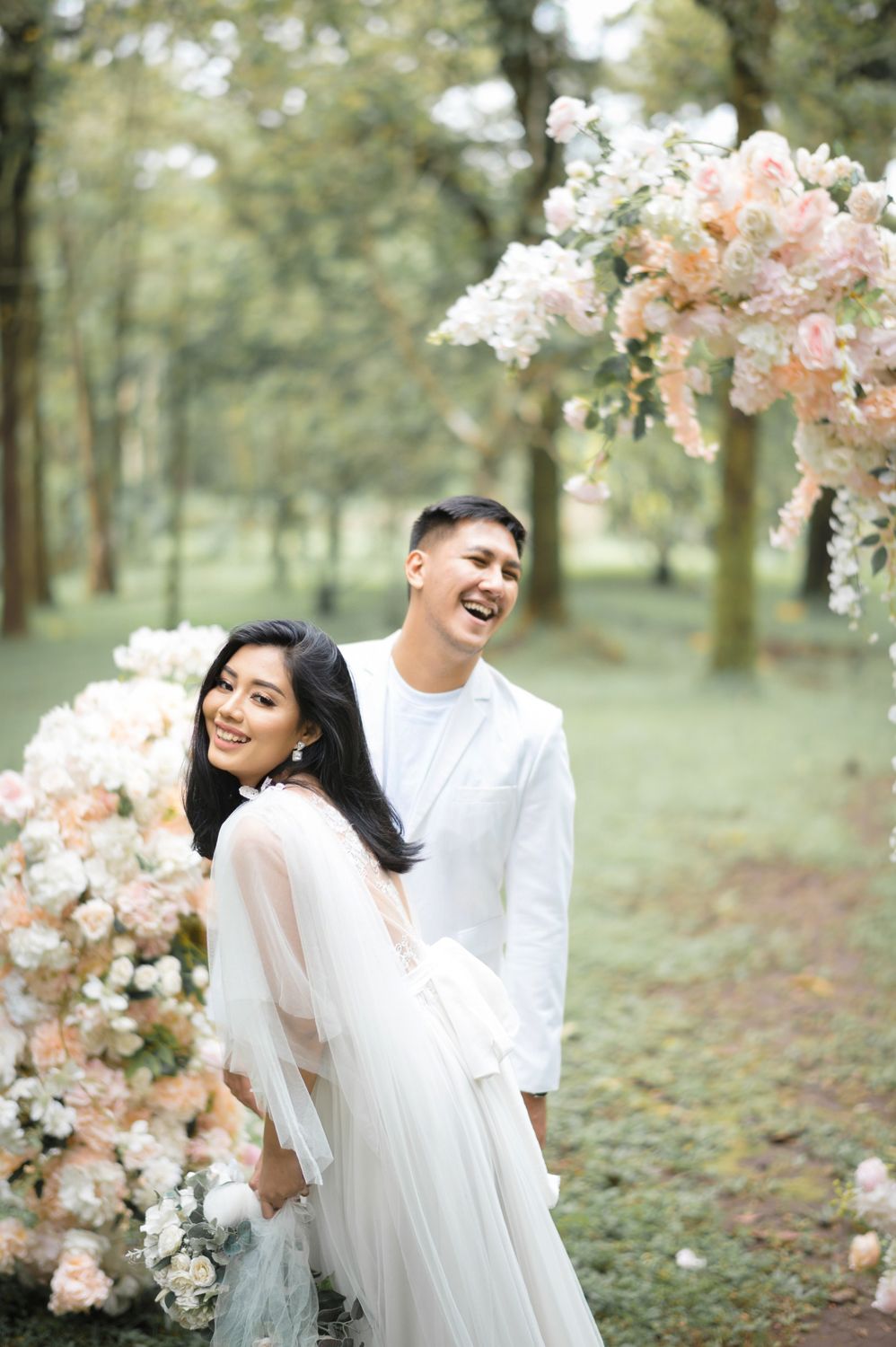 Couple in white attire laughing joyfully outdoors surrounded by blush pink floral arrangements in a lush green forest setting.