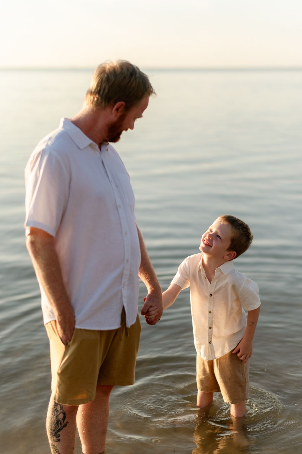 Father and son holding hands and smiling at each other while standing in calm ocean water at sunset.
