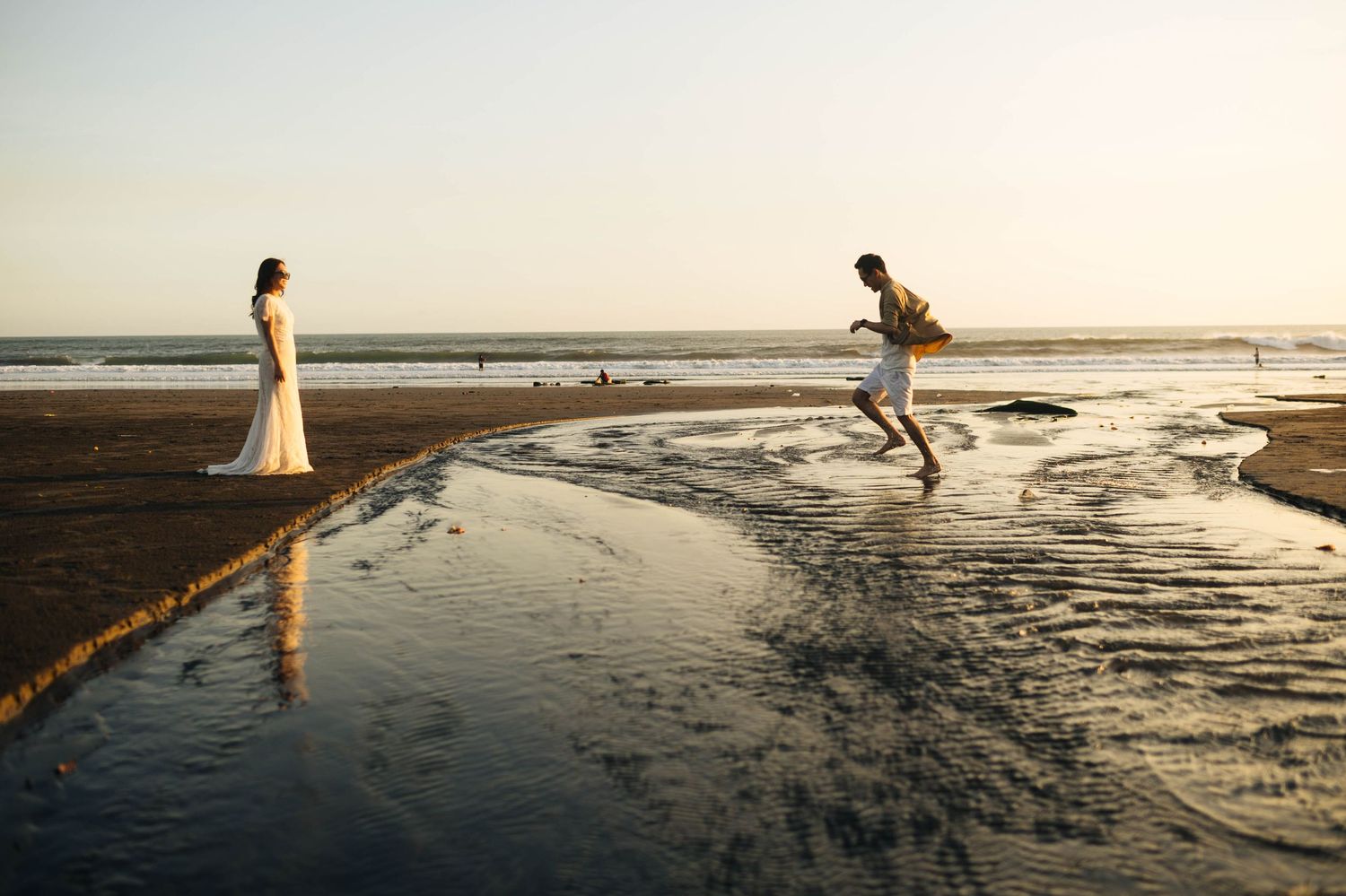 A groom leaps over water on a beach toward his bride in a wedding dress at sunset.