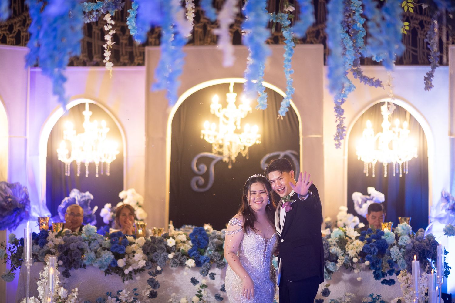 Couple smiles at wedding reception with blue floral arrangements, chandelier lighting, and elegant arched backdrop.