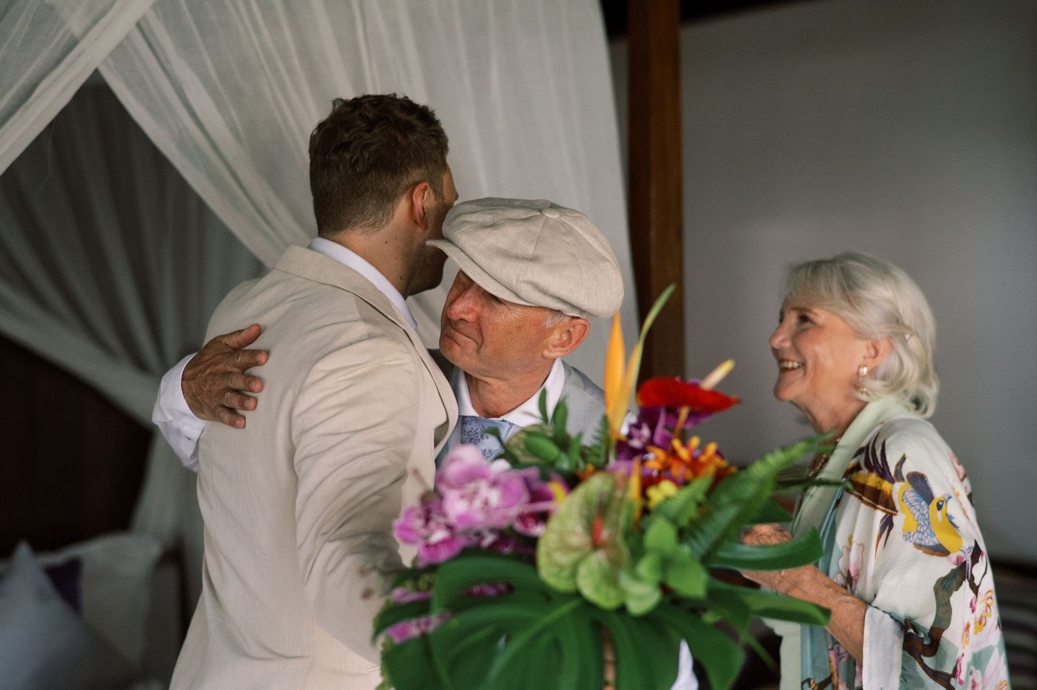 Groom embracing elderly man while woman smiles beside tropical flower arrangement.