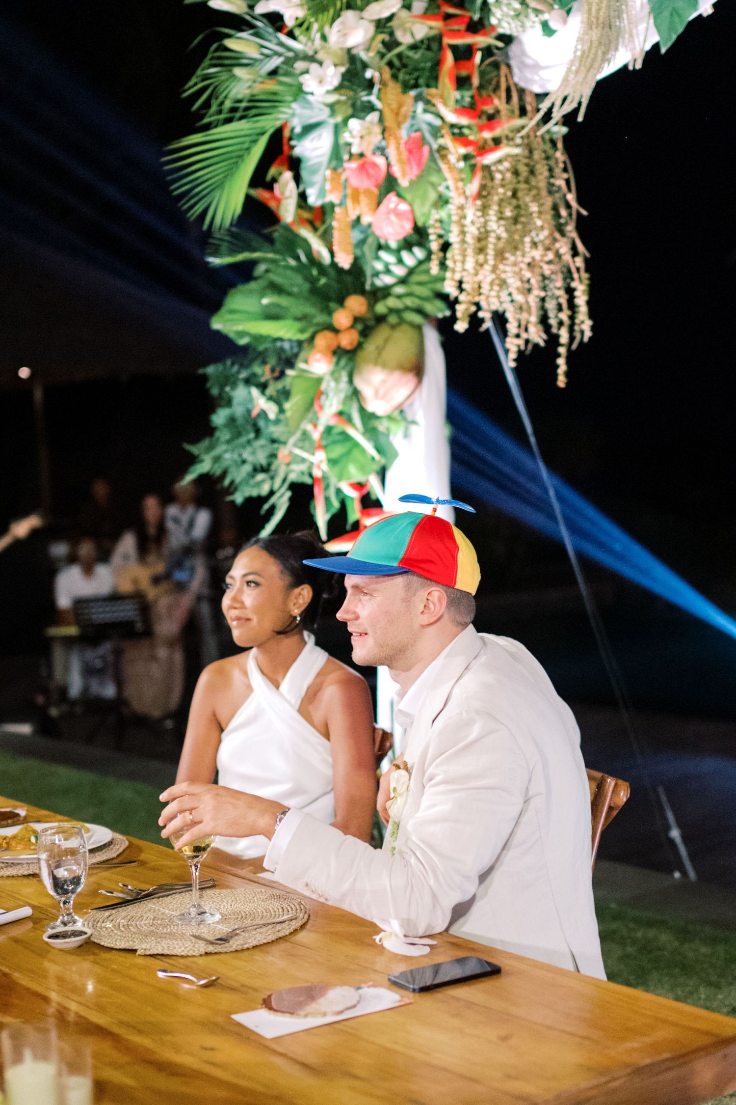Bride and groom sit at wedding reception table, groom wearing colorful propeller hat under tropical floral arrangement.