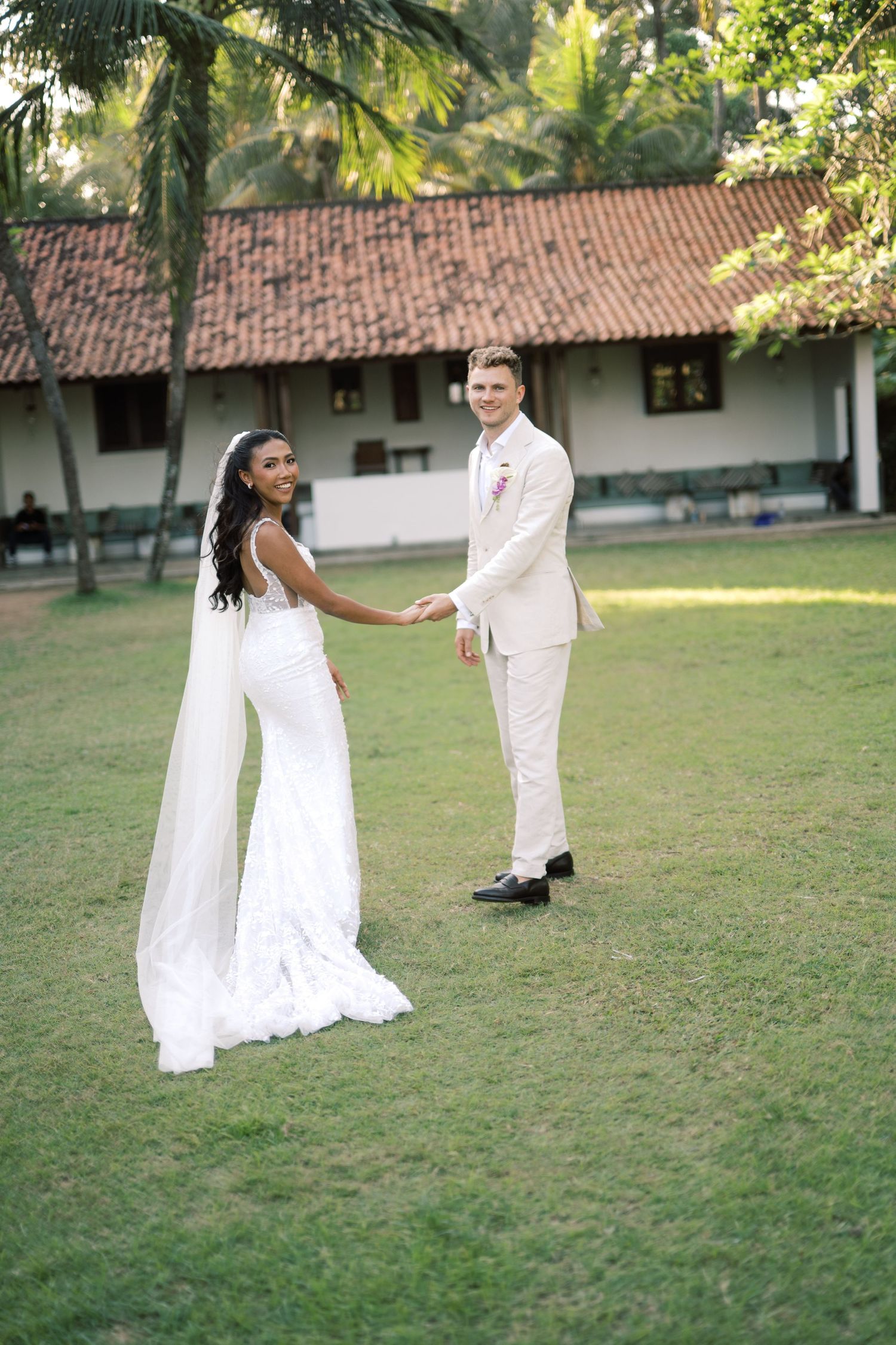 Bride in white cape gown and groom in beige suit hold hands on green lawn at tropical venue.