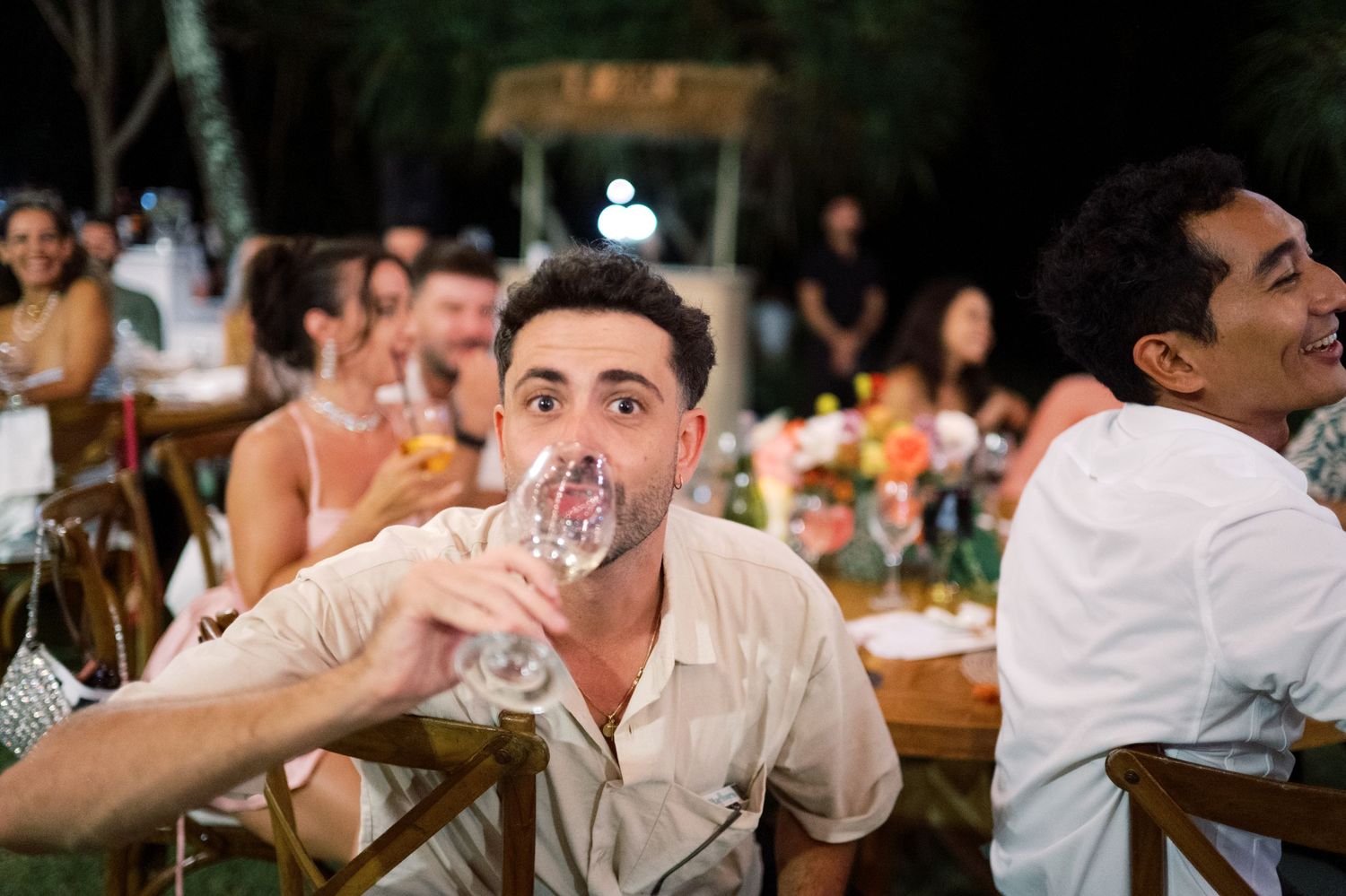 Wedding guest in white shirt drinks from a wine glass at a colorful outdoor tropical reception dinner.