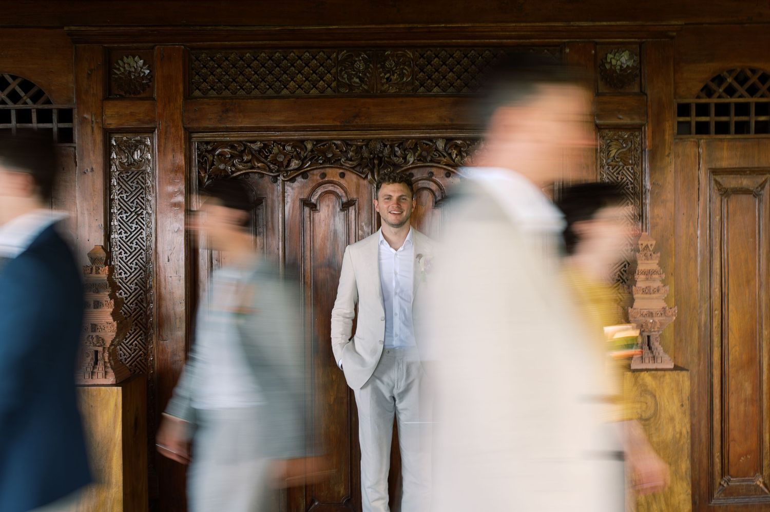 Groom in beige suit stands still as blurred wedding guests walk past ornate wooden doors.