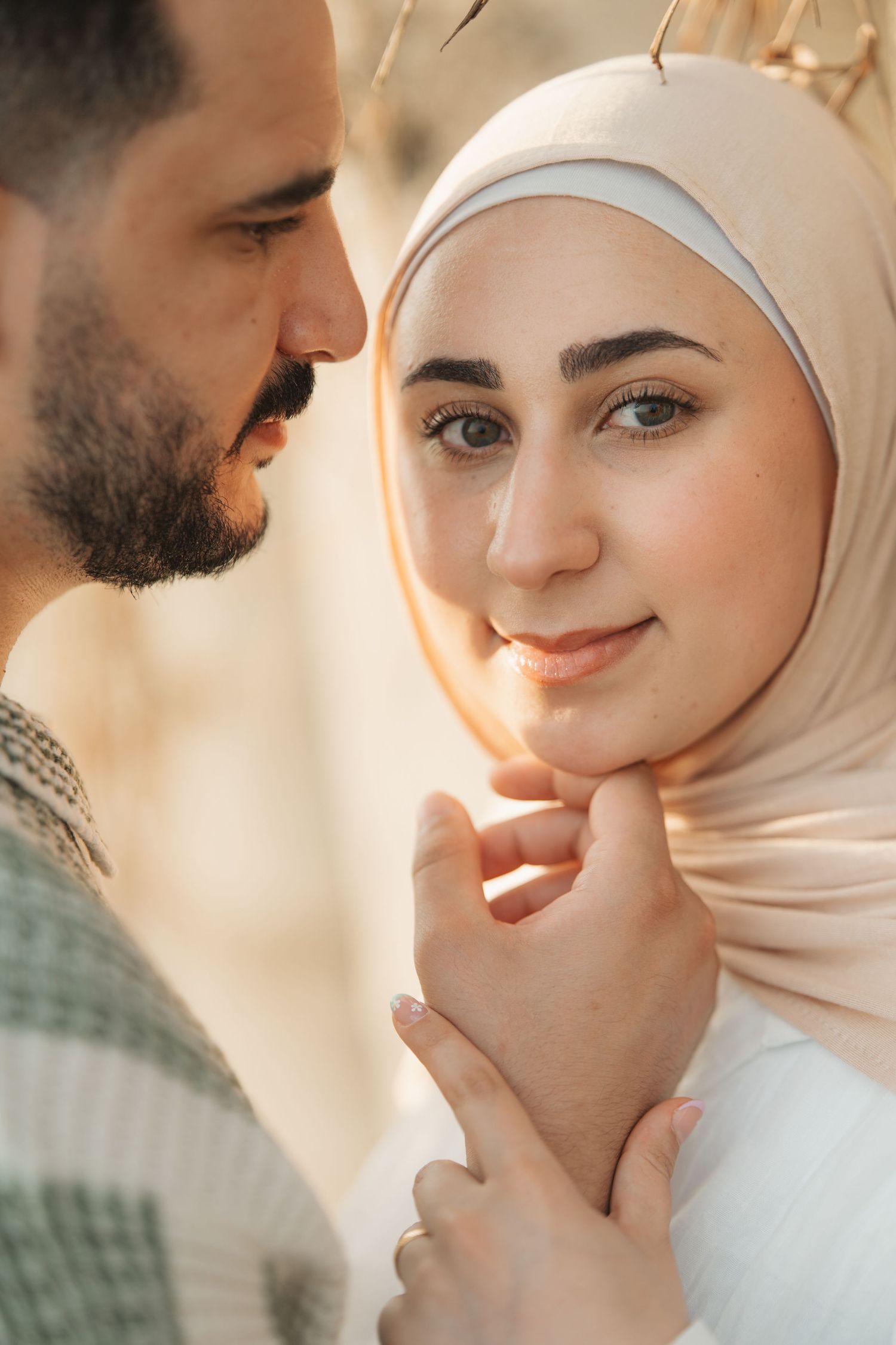 A Muslim couple shares a tender moment, the woman in a beige hijab smiling softly as the man gently holds her face.