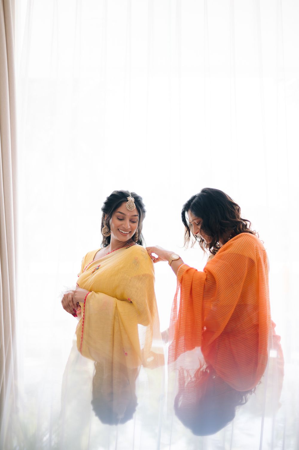 Bride in yellow saree smiles as woman in orange helps her dress near bright white curtains.