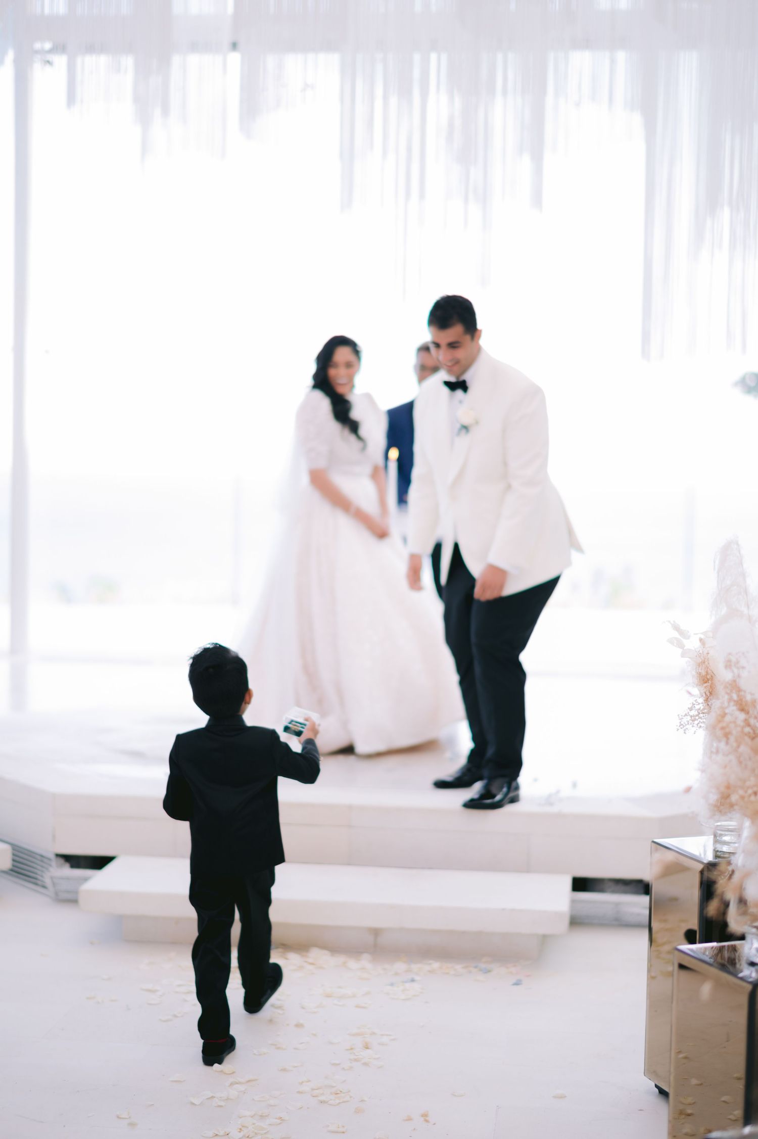 A young boy in a black suit watches a bride and groom at an elegant white wedding ceremony.