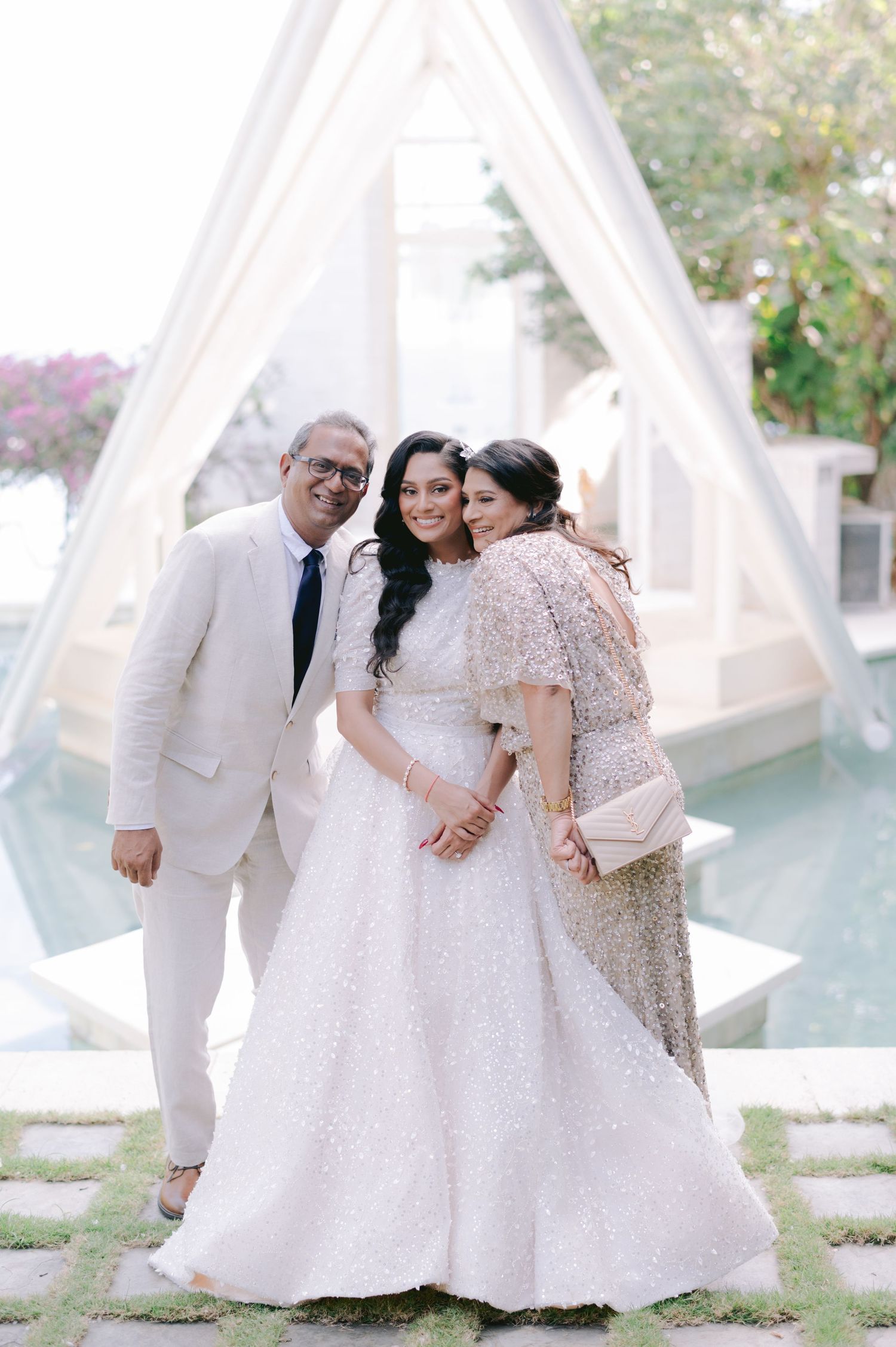 Bride in white lehenga poses with parents at elegant outdoor wedding venue with pool backdrop.