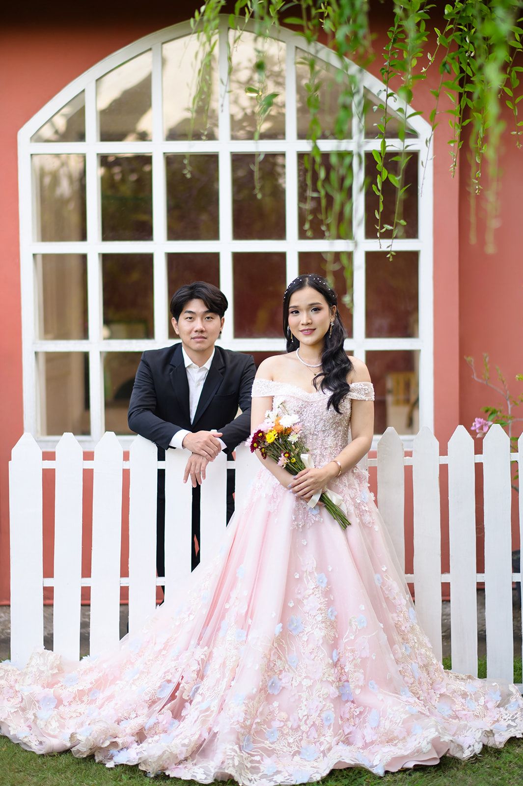 Couple posing in front of white picket fence, woman in pink floral gown holding bouquet, man in black suit.