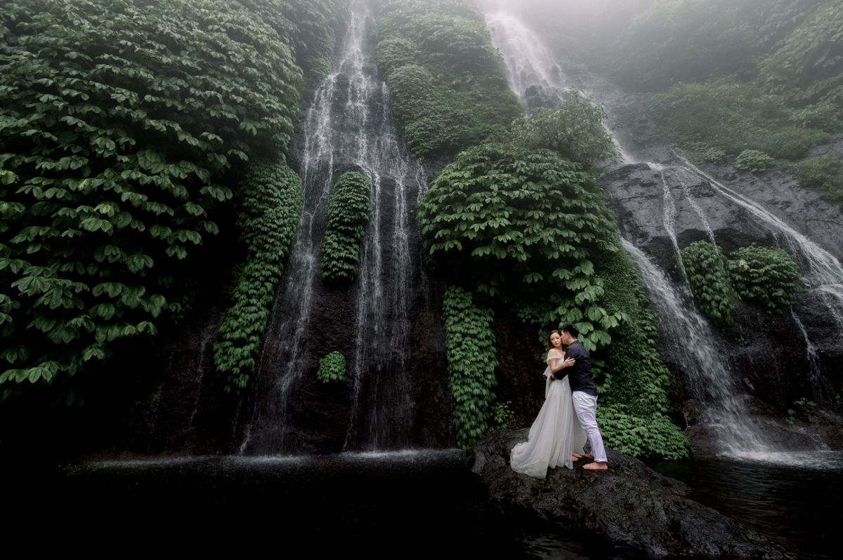 Couple embracing in wedding attire before a lush green waterfall in a misty tropical setting.