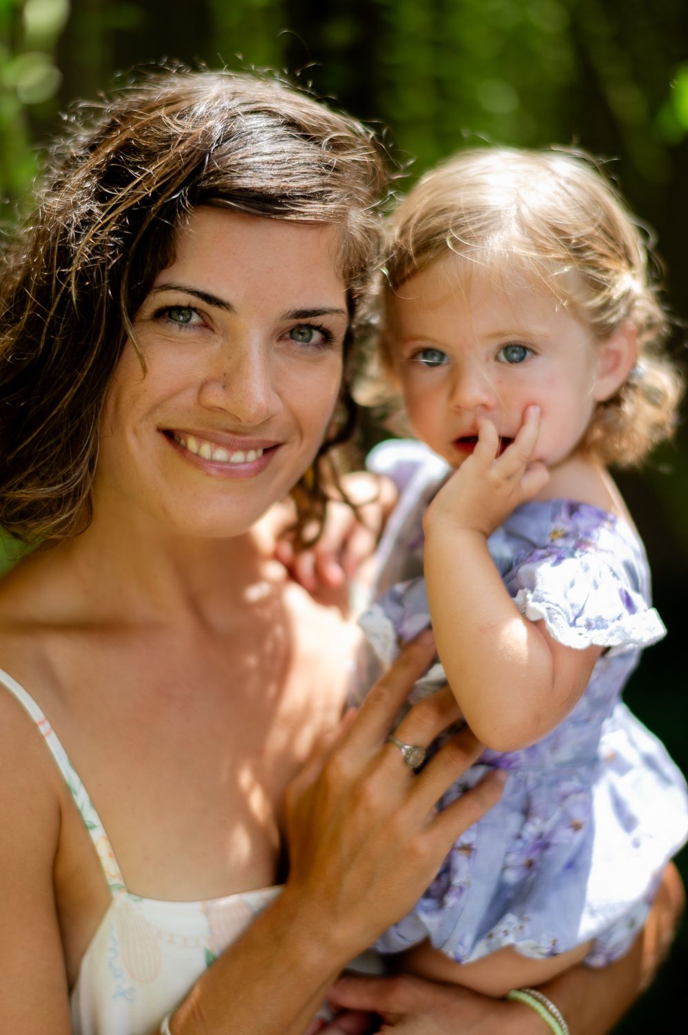 Smiling brunette woman holding a toddler girl in a purple floral dress outdoors in sunlight.
